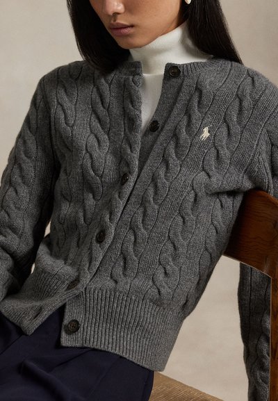 Nő szürke, fonott mintás gombos kardigánt visel fehér garbó felett, sötét nadrágban egy fa széken ülve.