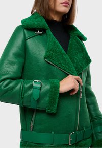 Zelená kožená bunda so stredným golierom a manžetami z ovčej vlny. Má strieborné zipsy a nastaviteľné bočné popruhy. Hladká textura a štruktúrovaný dizajn.