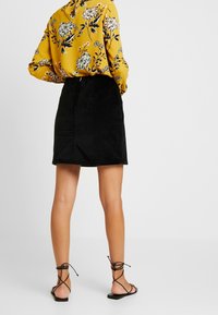 Woman wearing a black corduroy skirt, yellow floral long-sleeve blouse, and black lace-up sandals, standing on a plain light background.