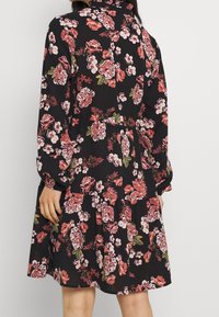 Květinové šaty z černé tkaniny s růžovými a bílými květy. Mají dlouhé rukávy, volánkový límec a rozšířenou sukni s přiléhavým pasem.