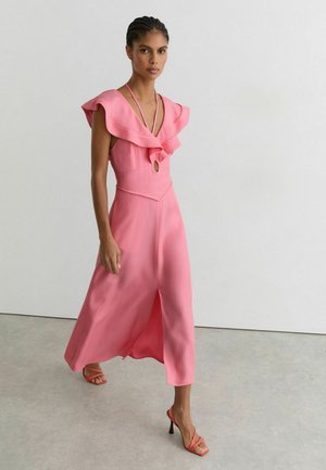 Kobieta idąca ubrana w różową sukienkę midi bez rękawów z marszczonym dekoltem i przednim rozcięciem, w połączeniu z koralowymi sandałkami na obcasie.