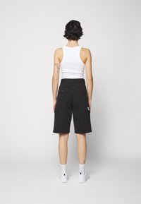 Shorts cargo noirs avec deux poches arrière et des poches latérales, associés à un débardeur blanc et des baskets blanches. Fond uni.