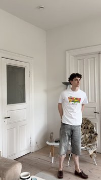 Hvit t-skjorte med et fargerikt grafisk design med teksten "Stay Fresh!" kombinert med grå denimshorts og brune sko. Innendørs setting.