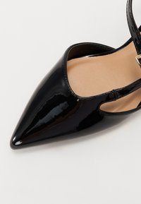 Črne lakirane slingback čevlje s koničastim nosom, izrezanimi stranmi in gladko bež notranjostjo. Zlati sponka.