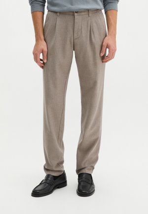 Man wearing beige pleated trousers and black leather loafers standing against a plain background.