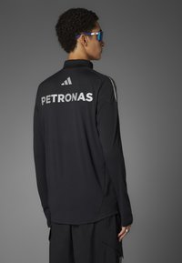 Sort langærmet skjorte med 'PETRONAS' logo i hvid på ryggen, med Adidas tre-stripes design på skuldrene. Glat tekstur.