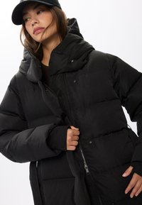 Giubbotto nero in materiale puffato con colletto alto, ispessimento termico, zip frontale con bottoni a pressione e polsini in maglia per una maggiore calore.