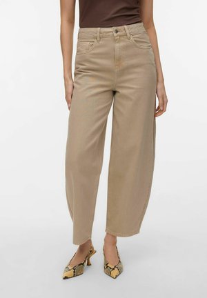 Femeie purtând pantaloni evazați bej cu talie înaltă, un top maro închis și pantofi cu toc ascuțiți din piele de șarpe galben, stând pe un fundal alb.