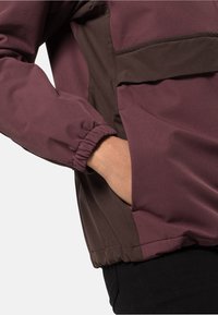 Veste bordeaux et marron avec poignets élastiques et une main dans la poche latérale, présentant un tissu doux et une coupe décontractée.