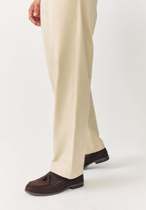 Uomo che indossa pantaloni beige e mocassini in pelle marrone scuro con nappine, in piedi davanti a uno sfondo bianco.
