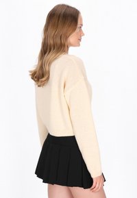 Crème, gebreide crop sweater met lange mouwen, gecombineerd met een zwarte geplooide mini rok. De outfit contrasteert in kleuren en texturen.