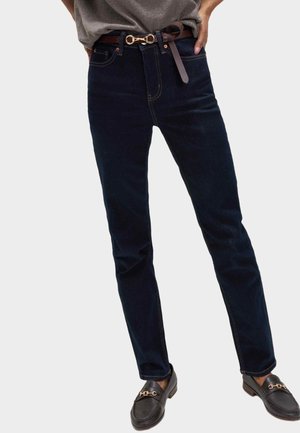 Persona que lleva jeans ajustados azul oscuro, cinturón de cuero marrón con hebilla dorada, camisa gris y mocasines marrón oscuro con detalle dorado.