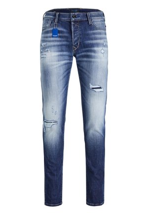 Homme portant un t-shirt blanc, un jean bleu déchiré et des baskets noires, assis sur un tabouret métallique industriel, bras croisés.