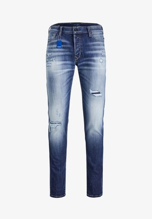 Mann trägt weißes T-Shirt, abgenutzte blaue Jeans und schwarze Sneakers, sitzt auf einem industriellen Metallhocker mit verschränkten Armen.