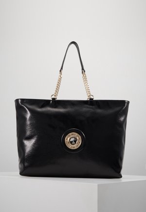 Czarna błyszcząca torba typu tote z uchwytami z złotego łańcuszka oraz okrągłą złotą emblematą z centralnym czarnym kamieniem i napisem marki.