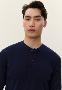 Fiatal férfi rövid sötét hajjal, tengerészkék hosszú ujjú henley felsőt visel, előre néz egy egyszerű bézs háttér előtt.