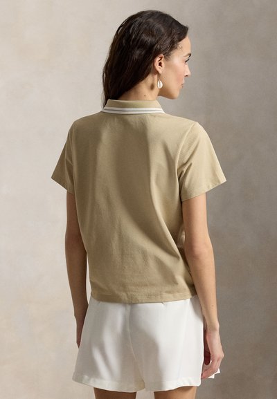 Žena s dlouhými tmavými vlasy, oblečená v béžovém polokošili s krátkým rukávem a bílých šortkách, otočená zády proti neutrálnímu pozadí.