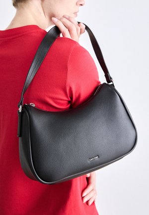 Osoba ubrana na czerwono trzyma czarną skórzaną torbę na ramię marki Calvin Klein z srebrnym zamkiem błyskawicznym i regulowanym paskiem na jednolitym białym tle.