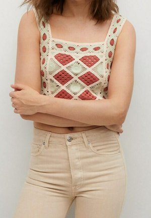 Mujer con un top corto de crochet sin mangas con patrones de diamantes rojos y crema y pantalones beige de cintura alta, con los brazos cruzados sobre el torso.