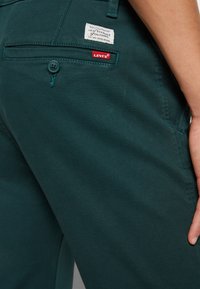 Gros plan sur la poche arrière d'un pantalon Levi's turquoise avec un bouton, une étiquette rouge Levi's, une étiquette de marque, et une main droite reposant sur le côté.
