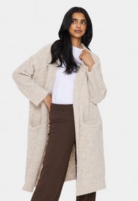 Beige stickad cardigan med långa ärmar, öppen front, två fickor och en mjuk textur, i kombination med bruna byxor och en vit skjorta.