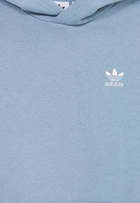 Světle modrá mikina s kapucí vyrobená z bavlněné směsi, s bílým logem trefoil Adidas na levé části hrudníku a texturovaným povrchem.