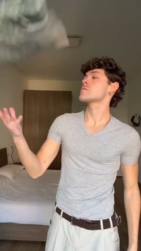 Mladý muž v šedém tričku hází vzorovaný kus látky v ložnici s postelí a skříní v pozadí.