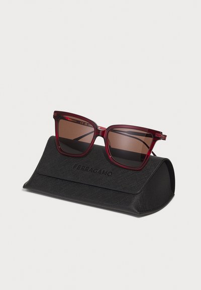 Sarkani kaķa acu saulesbrilles ar brūniem lēcu, ar caurspīdīgām apvalkām un zelta akcentiem, atpūšas melnā teksturētā maciņā ar uzrakstu "Ferragamo" iegravētu.