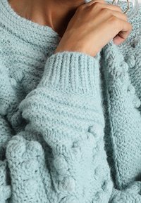 En närbild av en persons hand som håller ett mjukt, ljusblått stickat tröja med texturerade mönster och bollar på ärmarna.