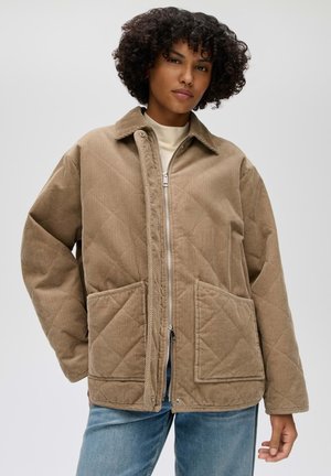 Femme aux cheveux bouclés portant une veste beige matelassée en velours côtelé avec des poches devant et une fermeture éclair, associée à un jean bleu clair.