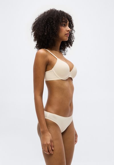 Γυναίκα με σγουρά μαλλιά, φοράει σετ μπεζ σουτιέν και εσώρουχα που ταιριάζουν, στέκεται προφίλ με φόντο απλό και φωτεινό.
