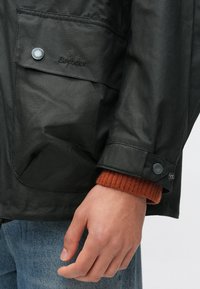 Schwarze gewachste Jacke mit einer Brusttasche und knöpfbaren Manschetten. Darunter ein kontrastierender oranger Strickpullover.