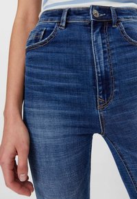 Plano detalle de una persona que lleva puesto unos vaqueros azul denim con el botón frontal y cremallera visibles, las costuras de los bolsillos y la mano relajada a un lado.