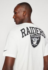 Camiseta blanca de manga corta con un gran texto y logo negro de "RAIDERS" en la parte posterior. Frontal liso con un logo negro de Nike en la manga.