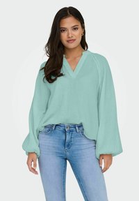 En lös, ljus tealblus med v-ringning, långa puffärmar och textilstruktur, kombinerad med figurnära blå jeans.