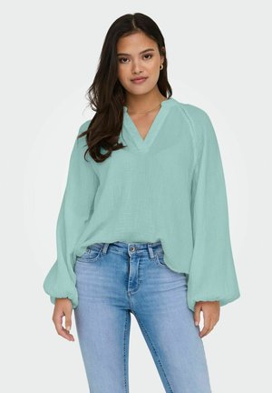 Eine lockere, hellblaue Bluse mit V-Ausschnitt, langen Puffärmeln und strukturiertem Stoff, kombiniert mit engen blauen Jeans.