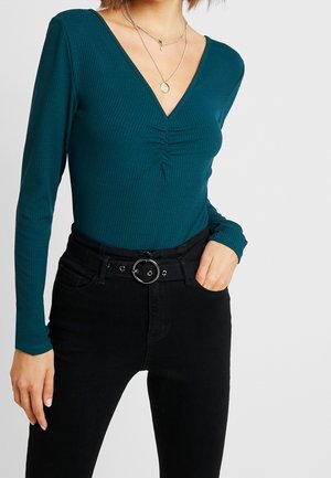 Kobieta ubrana w morski prążkowany top z długim rękawem i dekoltem w szpic z marszczeniem, czarne spodnie z wysokim stanem oraz warstwowe srebrne naszyjniki.