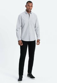 Vaaleanharmaa nappikauluspaita, jossa on rintatasku, yhdistettynä mustiin slim fit -housuihin ja mustiin lenkkareihin, seisoo yksinkertaista taustaa vasten.