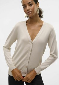 Gilet beige avec un col en V profond, des manches longues et cinq boutons marron. Texture douce et poignets et ourlet côtelés.