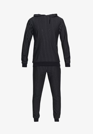 Mann trägt schwarzen Kapuzenpullover und Hose mit weißen Nadelstreifen, dazu weiße Turnschuhe, steht vor einem schlichten hellen Hintergrund.