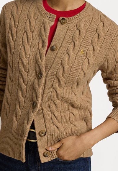 Cardigan bej tricotat cu model de cabluri, cu tivuri și manșete cu dantelă, prezentând nasturi din lemn și un mic logo brodat pe piept.