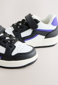 Children’s athletic shoes in white, black, and purple with laces and a Velcro strap on a neutral background.