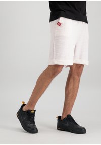 Shorts blancs texturés avec des ourlets retroussés et un patch logo rouge, associés à des Nike Air Force 1 noires avec des accents jaunes.