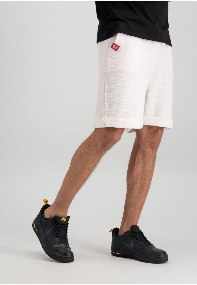 Shorts blancs texturés avec des ourlets retroussés et un patch logo rouge, associés à des Nike Air Force 1 noires avec des accents jaunes.