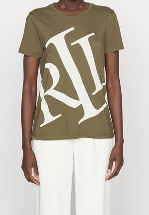 Persona de piel oscura que lleva puesta una camiseta verde oliva con grandes letras blancas superpuestas y pantalones blancos, de pie contra un fondo liso.