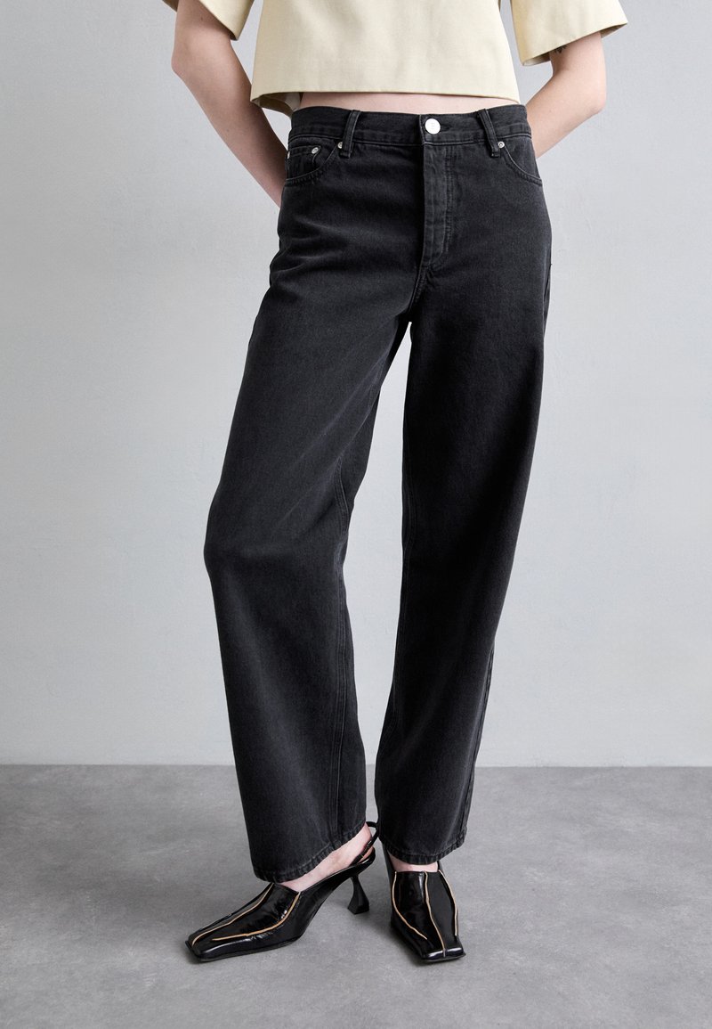 Black denim wide-leg jeans with a high waist, featuring front pockets and a button closure. Paired with black patent leather heeled shoes.