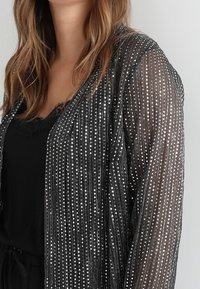 Sort kimono med pailletdetaljer i et let, gennemsigtigt stof. Lodrette striber af sølvpailetter skaber en tekstureret, reflekterende effekt.