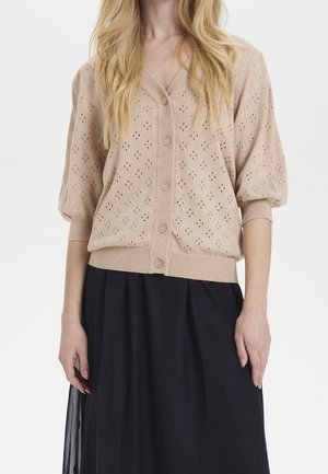 Frau trägt eine hellbeige gestrickte, geknöpfte Strickjacke mit Puffärmeln und einen schwarzen plissierten Rock vor weißem Hintergrund.