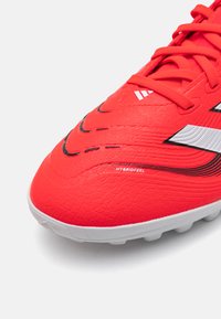 Talon de football rouge avec une surface texturée, doté d'accents noirs et de rayures blanches. Comprend le logo "HYBRIDFEEL" sur le côté.