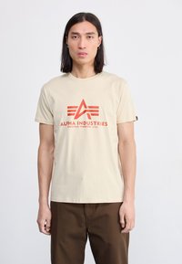 Beige cotton t-shirt featuring a bold red “Alpha Industries” logo with an emblematic design. Short sleeves and a classic crew neckline.